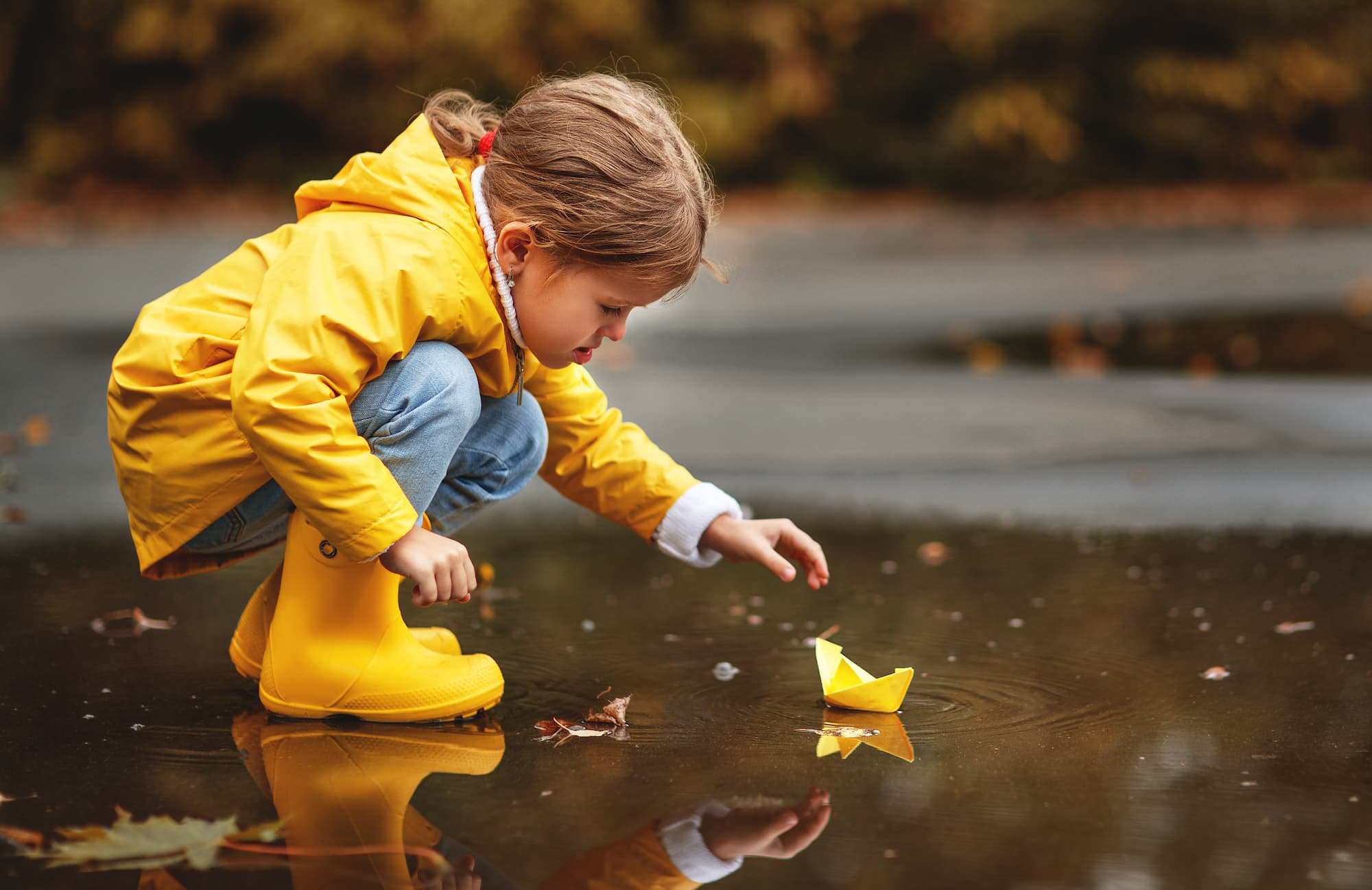 A child in a bright yellow raincoat playing with their paper boat in a puddle