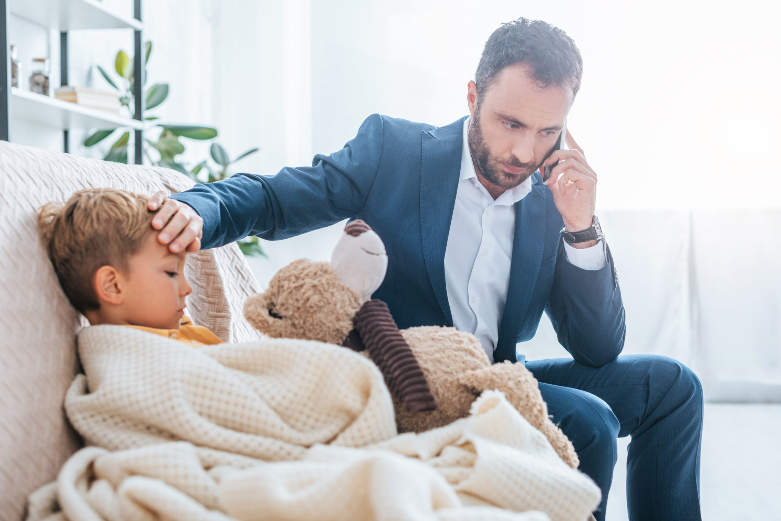 worried father touching forehead of sick son while talking on smartphone