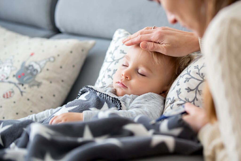 Sick child, toddler boy lying on the couch in living room with a fever, mother checking their temperature
